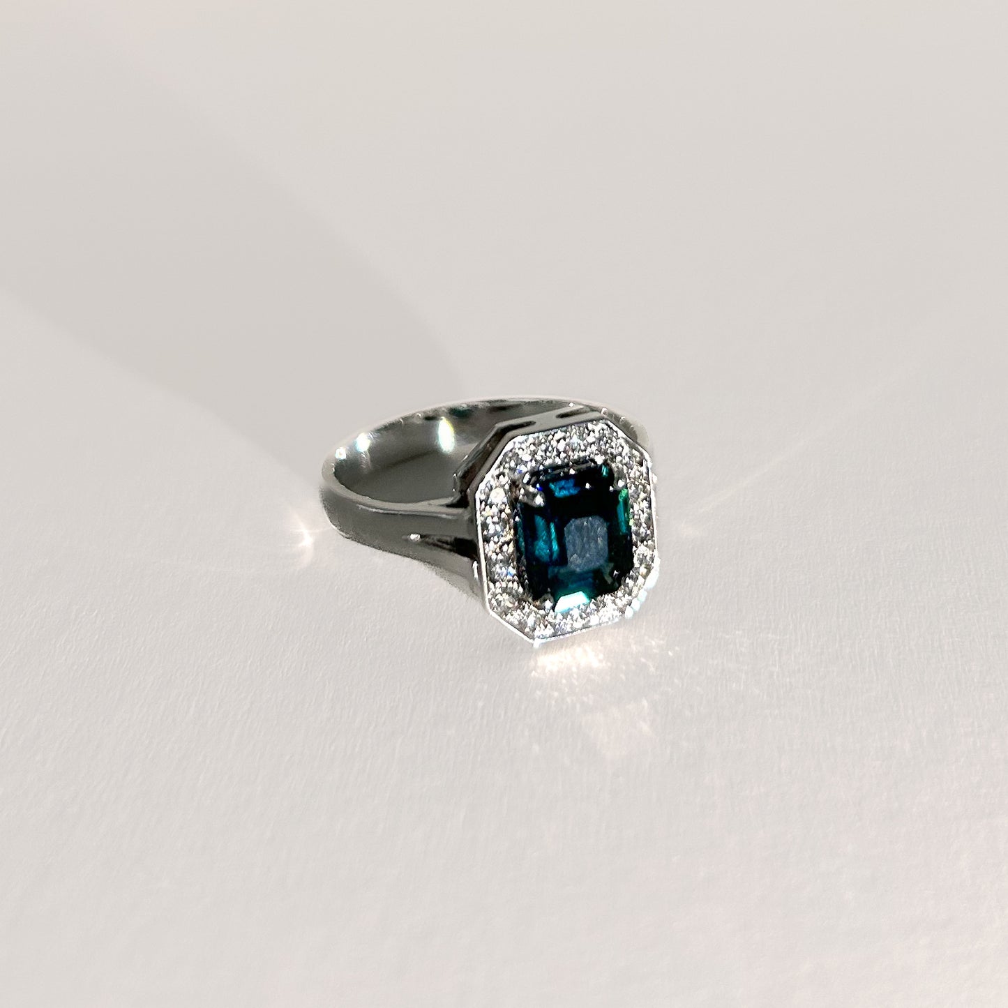 Art Deco-style 18K white gold ring featuring a 1.56ct emerald-cut unheated teal sapphire, surrounded by a halo of diamonds, offering a bold, timeless statement.