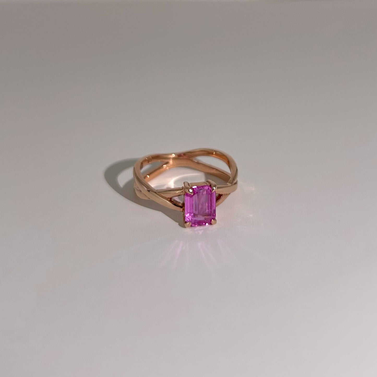 Vibrant 1.1ct Emerald cut Pink Sapphire set in 18K yellow gold, with a unique criss-cross band design for a modern and playful engagement ring.