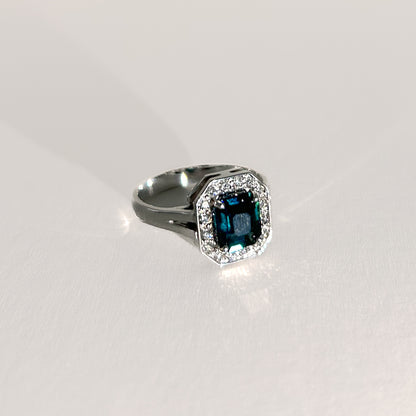 Art Deco-style 18K white gold ring featuring a 1.56ct emerald-cut unheated teal sapphire, surrounded by a halo of diamonds, offering a bold, timeless statement.