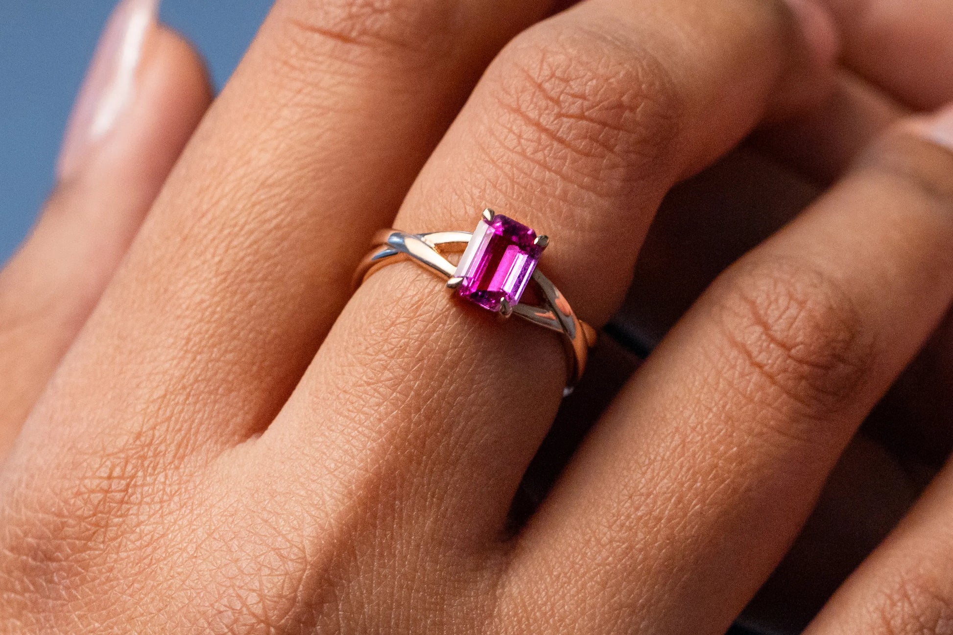 Custom 18K yellow gold engagement ring featuring a 1.1ct unheated Emerald cut Pink Sapphire from Sri Lanka, with criss-cross bands for a unique and modern design.