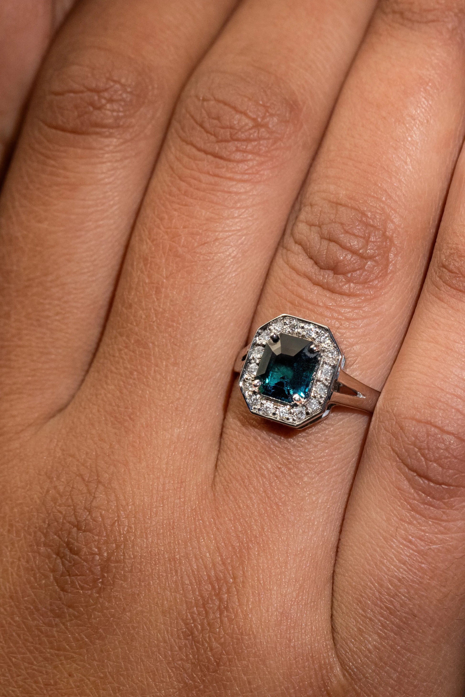 Close-up of 18K white gold ring with 1.56ct unheated emerald-cut teal sapphire, encircled by a halo of 0.12ct diamonds, inspired by Art Deco glamour.