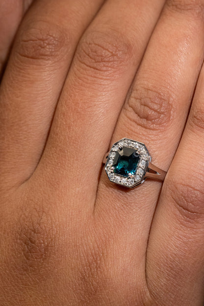 Close-up of 18K white gold ring with 1.56ct unheated emerald-cut teal sapphire, encircled by a halo of 0.12ct diamonds, inspired by Art Deco glamour.